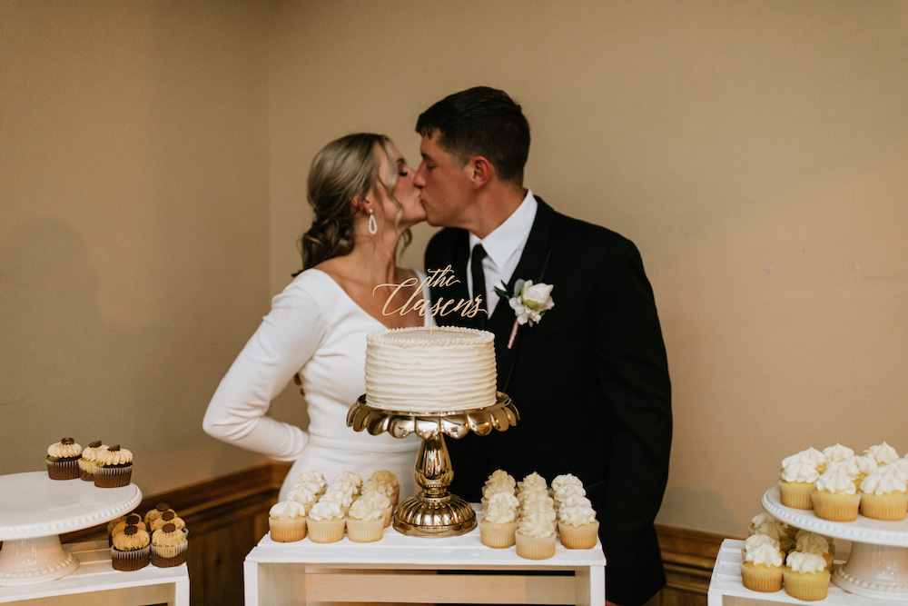 Bride and groom kissing in front of their white buttercream wedding cake on a gold stand.