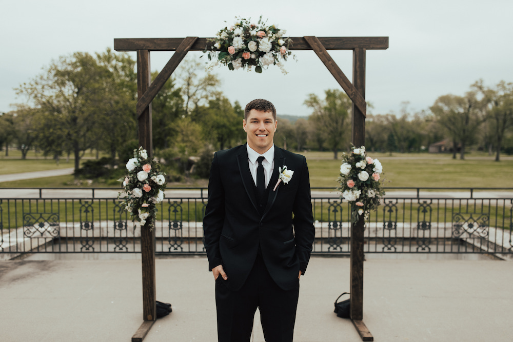 Elegant groom photo standing in front of the wedding arch wearing a black tux and black tie with a white boutonniere. 