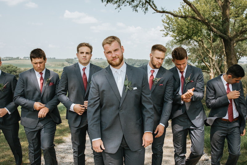 Groom and groomsman wearing dark grey suits and red and white ties. 